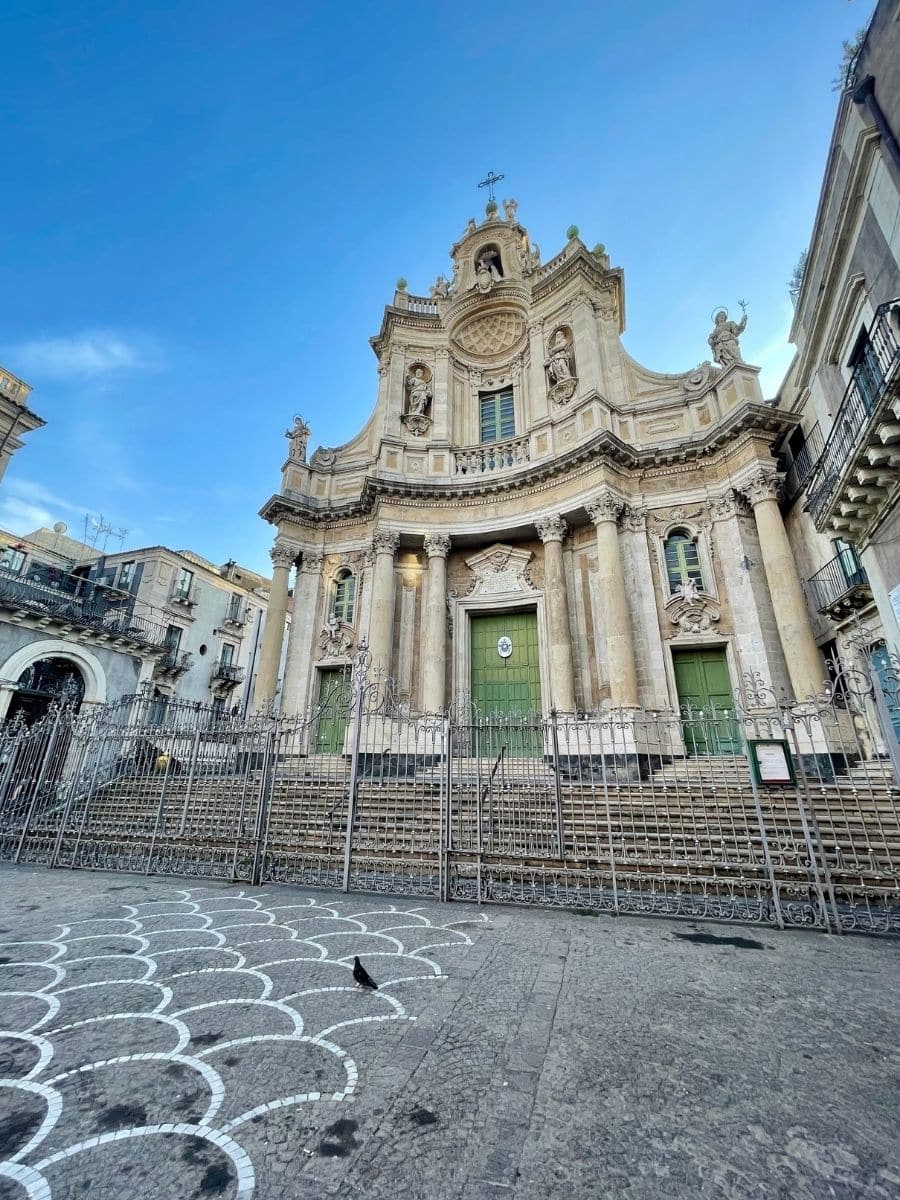 A view of the Basilica della Collegiata, a historic church in Catania, Sicily, showcasing its baroque architecture, statues, and the piazza in front with its distinctive paving pattern.