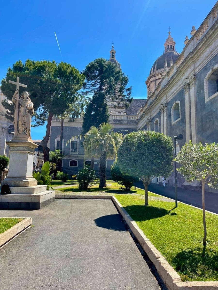 A peaceful garden next to a historic church in Catania, Italy, featuring a statue of a religious figure holding a cross. The manicured greenery, palm trees, and classic architecture, including the domes and ornate details of the church, create a serene and picturesque atmosphere under a bright blue sky.