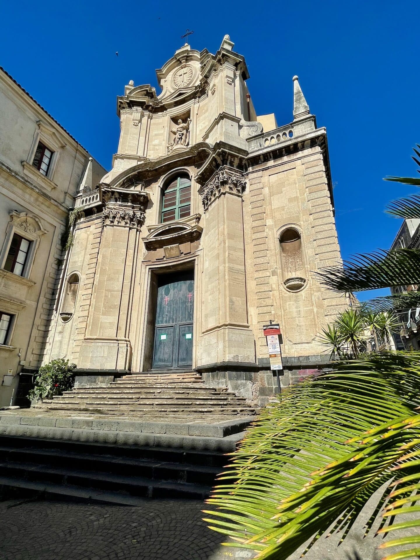 A baroque church facade in Catania with a clock and statues, viewed from below.
