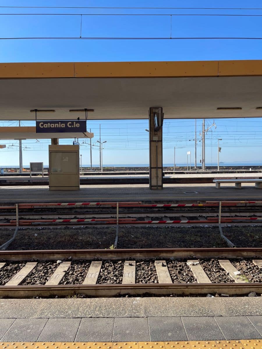 iew of Catania Centrale train station platform with a clear blue sky. The station sign 'Catania C.le' is visible above the empty tracks and platform, highlighting the calm and quiet of the station.