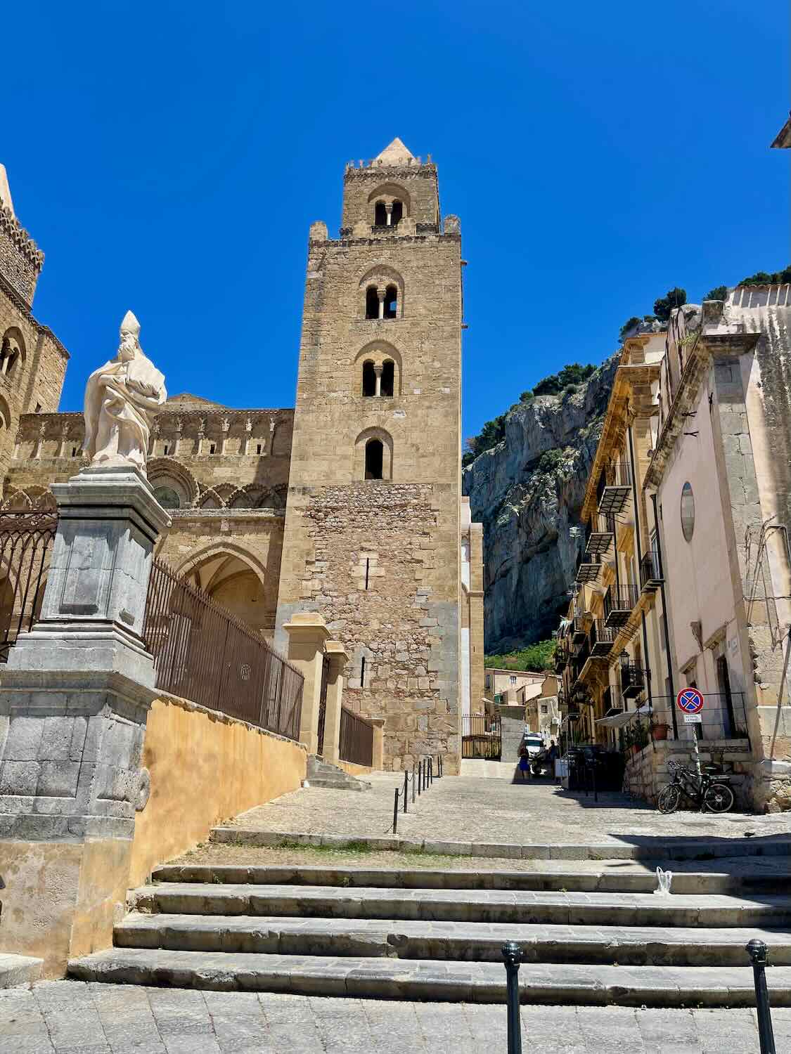 A historic tower in Cefalù, Sicily, rises against a clear blue sky. A statue stands on a pedestal in the foreground, and stone steps lead up to the tower. The scene includes traditional buildings and a rocky hillside.