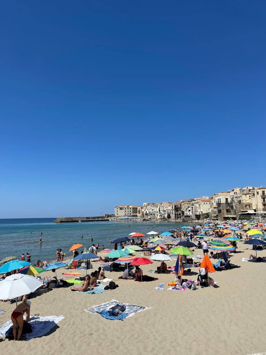 A lively beach in Cefalù, Sicily, filled with colorful umbrellas and sunbathers on golden sand. The calm, clear blue waters of the Mediterranean stretch towards the horizon, where people are seen swimming and wading. In the background, historic buildings with warm-toned facades and terracotta roofs line the waterfront, adding to the picturesque charm of the scene. The sky is a brilliant, cloudless blue, emphasizing the perfect beach day atmosphere.