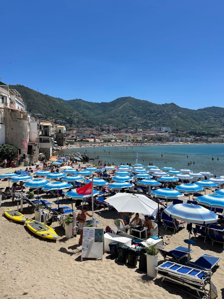 A lively beach in Cefalù, Sicily, with rows of blue and white umbrellas shading sunbathers on golden sand. The turquoise waters of the Mediterranean stretch out to meet a backdrop of lush green mountains and a picturesque coastal town. Beachgoers relax under the umbrellas, swim in the sea, or enjoy meals at seaside tables. Yellow paddleboards rest on the sand near a red flag, while a white signboard stands at the entrance to the beach area. The scene captures the vibrant atmosphere of a Sicilian summer by the sea.