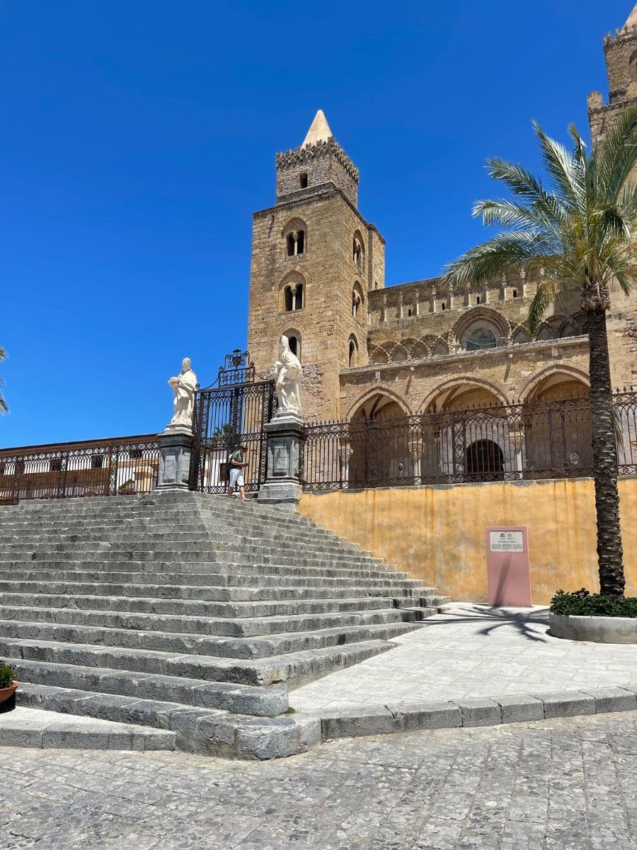 A majestic medieval cathedral with two imposing bell towers rises against a brilliant blue sky. A grand stone staircase leads up to the entrance, flanked by two statues on pedestals. The cathedral's intricate stonework and arched entrances showcase its historic architectural beauty. A tall palm tree stands to the right, adding a touch of greenery to the scene. A single visitor is seen walking up the stairs, giving a sense of scale to the structure. The warm-toned walls contrast beautifully with the cool, textured stone steps and cobblestone pavement in the foreground.