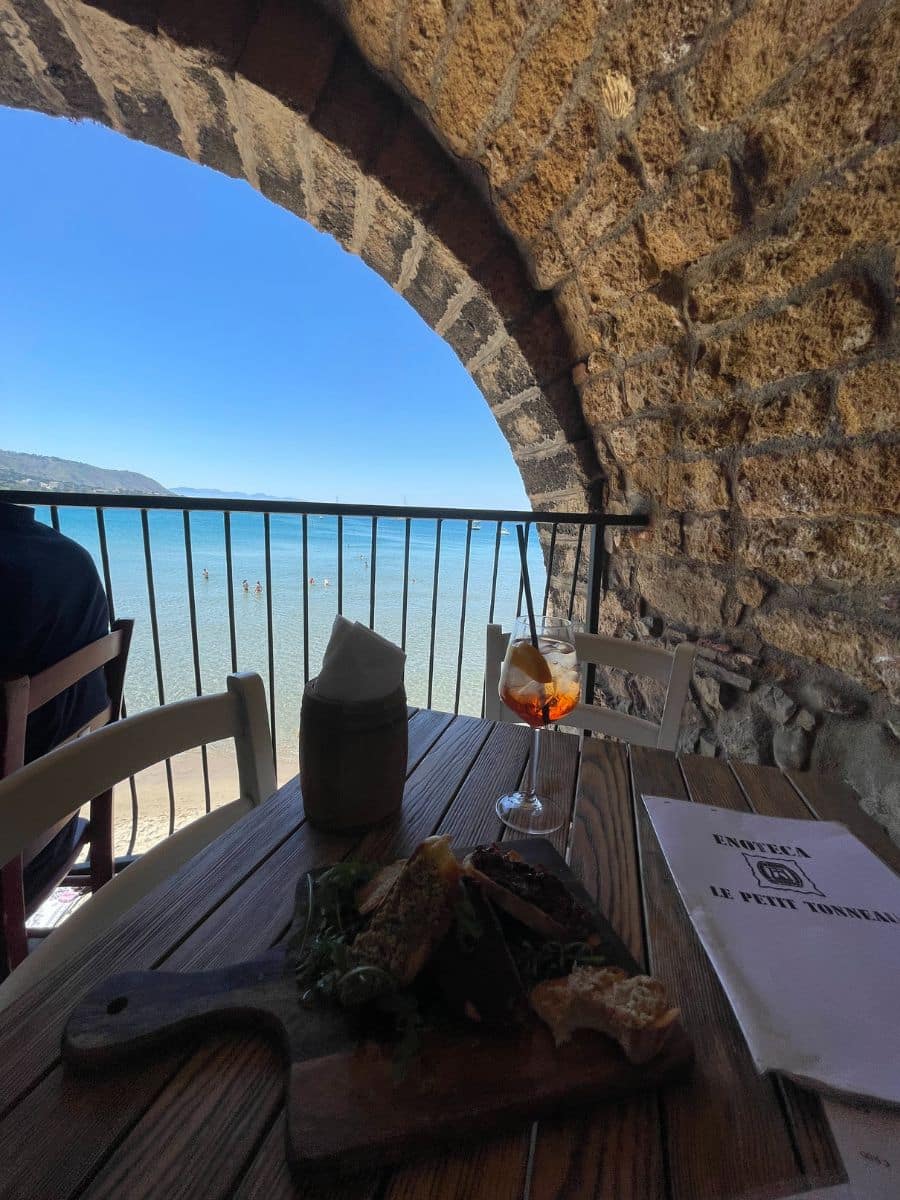 A rustic seaside dining scene framed by an ancient stone archway. The wooden table holds a charcuterie board with fresh bread, greens, and various toppings, along with a glass of Aperol spritz. A menu labeled 'Enoteca Le Petit Tonneau' is placed beside the meal. Through the arched opening, a breathtaking view of the calm blue sea with beachgoers and distant hills is visible. The contrast between the historic stone and the serene coastal setting creates a warm and inviting ambiance.