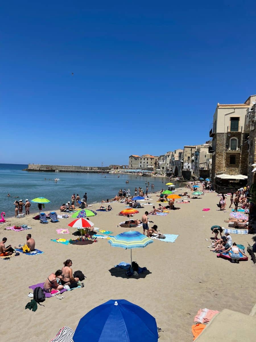 A lively beach in Cefalù, Sicily, with golden sand filled with colorful umbrellas, sunbathers, and beachgoers enjoying the Mediterranean sun. The calm, clear blue sea stretches towards the horizon, while historic stone buildings with charming balconies and shuttered windows line the coastline, giving the scene a picturesque, old-world charm. The sky is a vibrant shade of blue, completing the idyllic summer atmosphere.