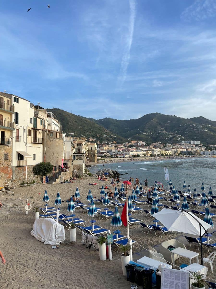 A scenic beach in Cefalù, Sicily, with rows of blue sun loungers and umbrellas on the sand, surrounded by historic buildings and a mountainous backdrop, as people enjoy the shoreline and the sea.