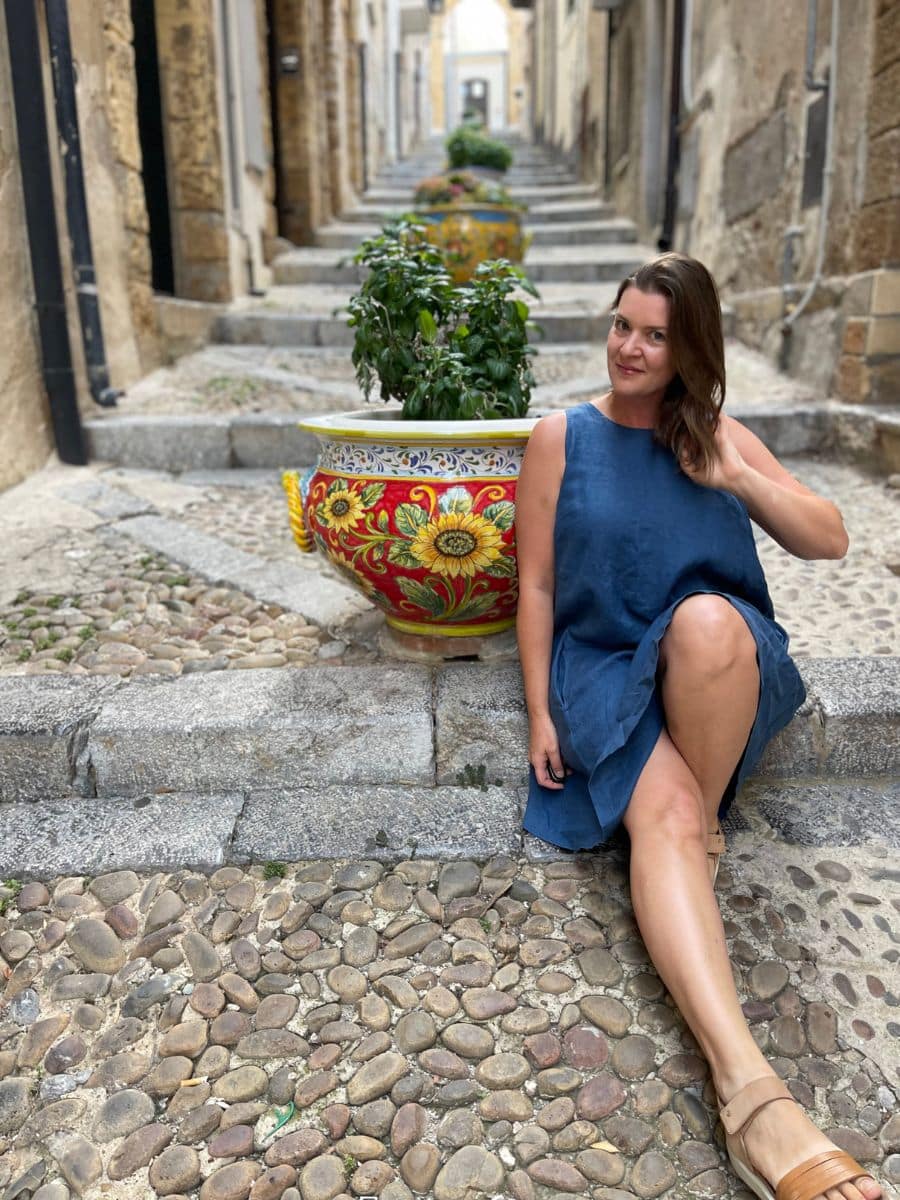 Melissa in a blue linen dress sits on a cobblestone street in Cefalù, Italy, resting against a stone step. Behind her, a staircase lined with ornate, hand-painted Sicilian ceramic pots filled with lush green plants adds vibrant color to the historic alleyway. The weathered stone walls and narrow passage create an old-world charm, capturing the essence of this picturesque Sicilian town.