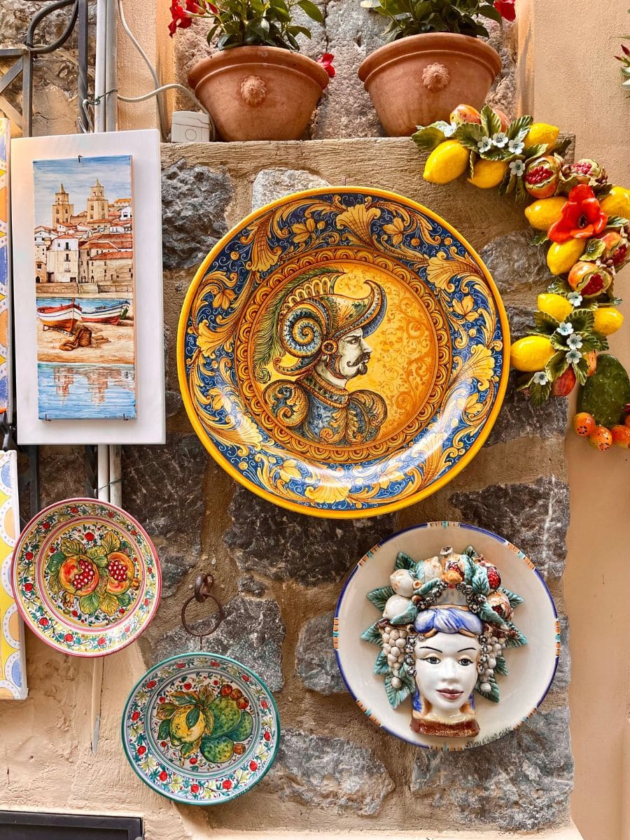 A display of traditional Sicilian ceramic plates mounted on a rustic stone wall in Cefalù, Sicily. The centerpiece is a large, ornate plate featuring a Moorish warrior, surrounded by smaller hand-painted plates with vibrant fruit motifs. A decorative wreath of lemons and pomegranates adds a Mediterranean charm, alongside a sculpted ceramic face adorned with flowers and fruit.