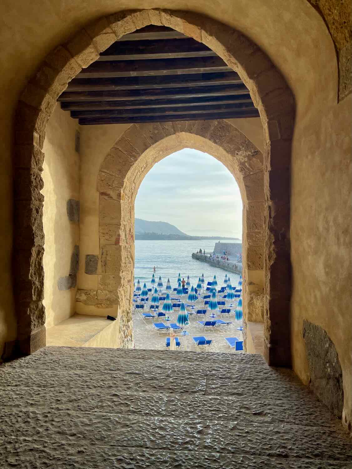 A view of a beach in Cefalù, Sicily, seen through a stone archway. Blue beach umbrellas and sunbeds are arranged on the sandy shore, with the sea and a distant pier visible in the background.