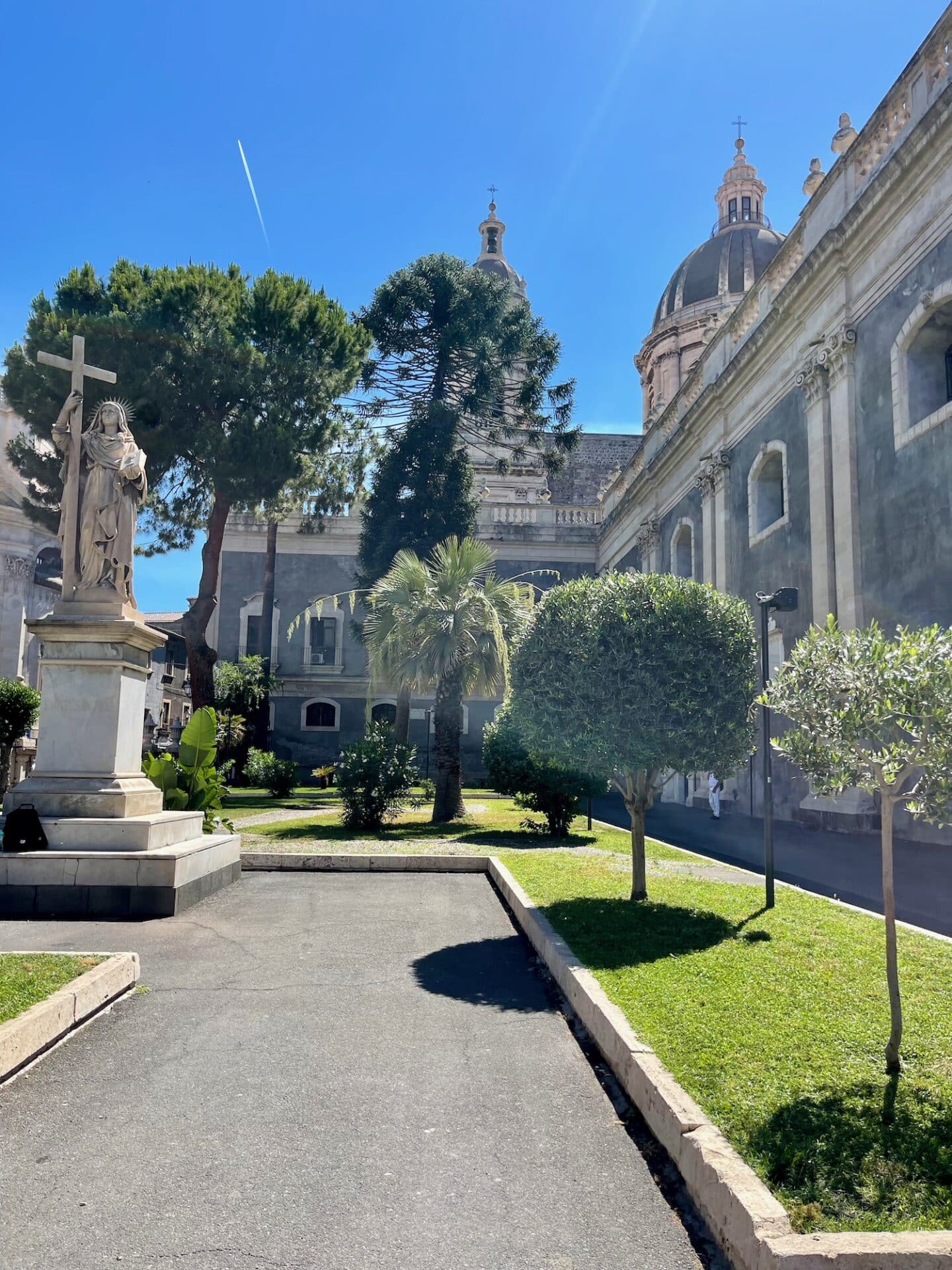 A courtyard in Catania featuring a statue with a cross, lush greenery, and domed buildings in the background.