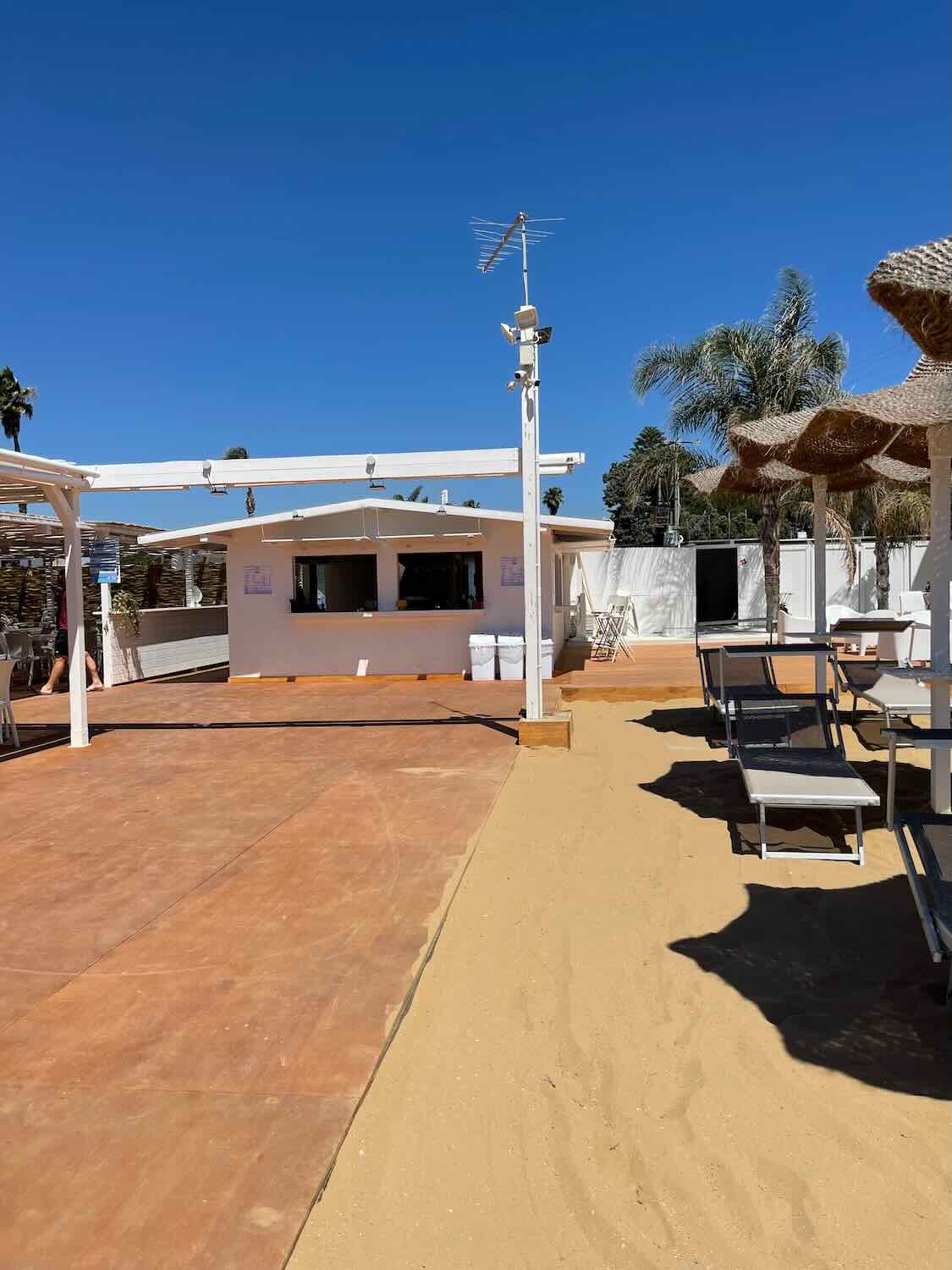 An outdoor club area with a sandy floor and several tables and chairs arranged under a white canopy. The sky is clear and blue, and there are palm trees in the background.