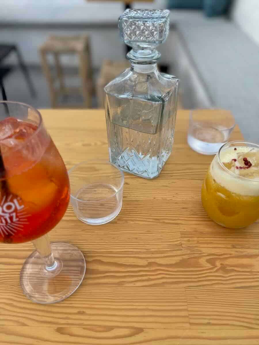 The image shows two drinks on a wooden table: an Aperol Spritz on the left and a frothy orange drink on the right, garnished with red berries. A crystal water decanter is placed between them, adding an elegant touch. The setting appears casual and outdoors.