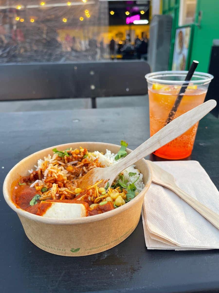 A close-up of a takeaway meal in a biodegradable bowl, featuring white rice topped with spicy tomato-based curry, paneer cheese, crispy chickpeas, and fresh cilantro. A wooden fork rests in the bowl. Next to it, a plastic cup with a refreshing orange-red beverage, garnished with lemon slices and a black straw, sits on a black table. A napkin and extra wooden spoon are nearby. In the blurred background, an outdoor dining area with string lights and people enjoying their meals creates a lively atmosphere.