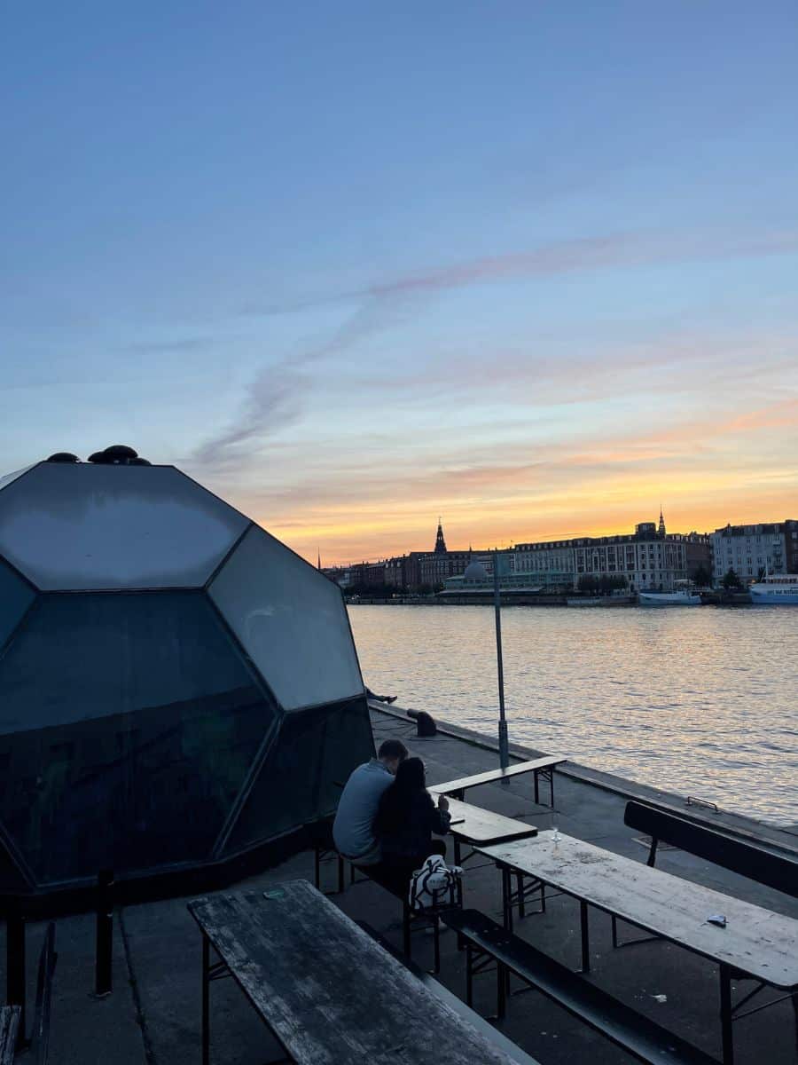 A waterfront setting at sunset with a sky painted in hues of orange, pink, and blue. A large geodesic dome structure with dark reflective panels stands on the left. A couple sits at a worn wooden bench, facing the water, engaged in conversation. The calm water reflects the fading sunlight, with a cityscape of historic buildings and church spires lining the opposite shore. Boats are moored along the waterfront, and the scene has a peaceful, intimate atmosphere.