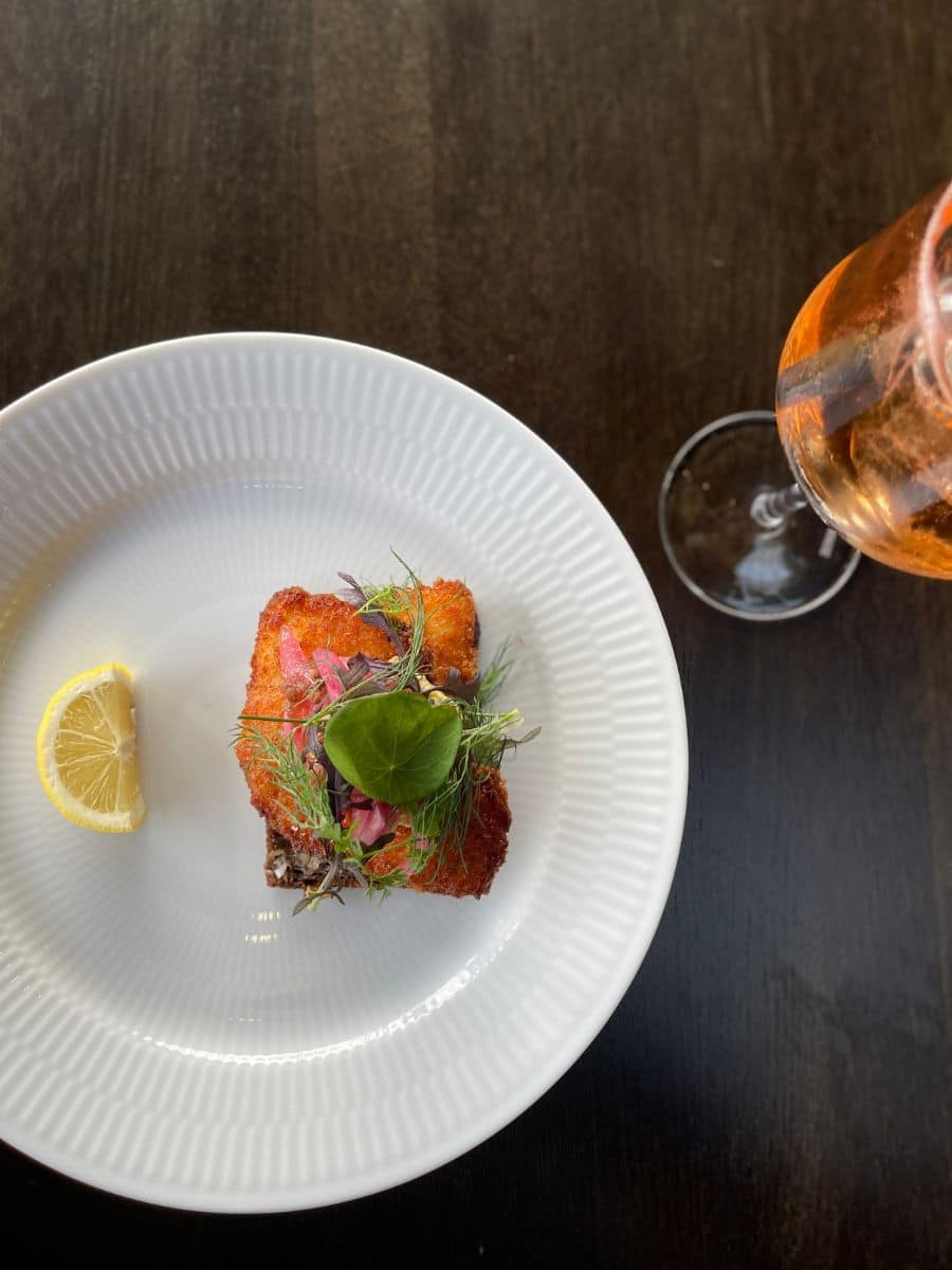 A crispy golden-brown piece of fried fish served on a slice of dark rye bread, garnished with fresh dill, pickled red onions, and a green leaf. A wedge of lemon sits on the side of a white textured plate. To the right, a glass of sparkling rosé wine is partially visible on a dark wooden table.