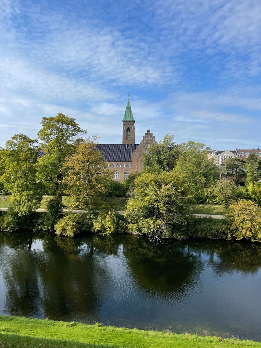 A scenic view of a historic brick building with a green spire, surrounded by lush trees, reflecting in the calm waters of a nearby river under a bright blue sky with scattered clouds in Copenhagen.