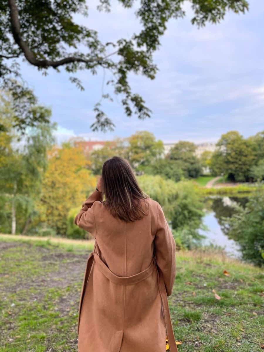 Back view of a woman in a beige coat overlooking a picturesque lake surrounded by dense foliage in a Copenhagen park, highlighting the city's natural beauty and peaceful environment.