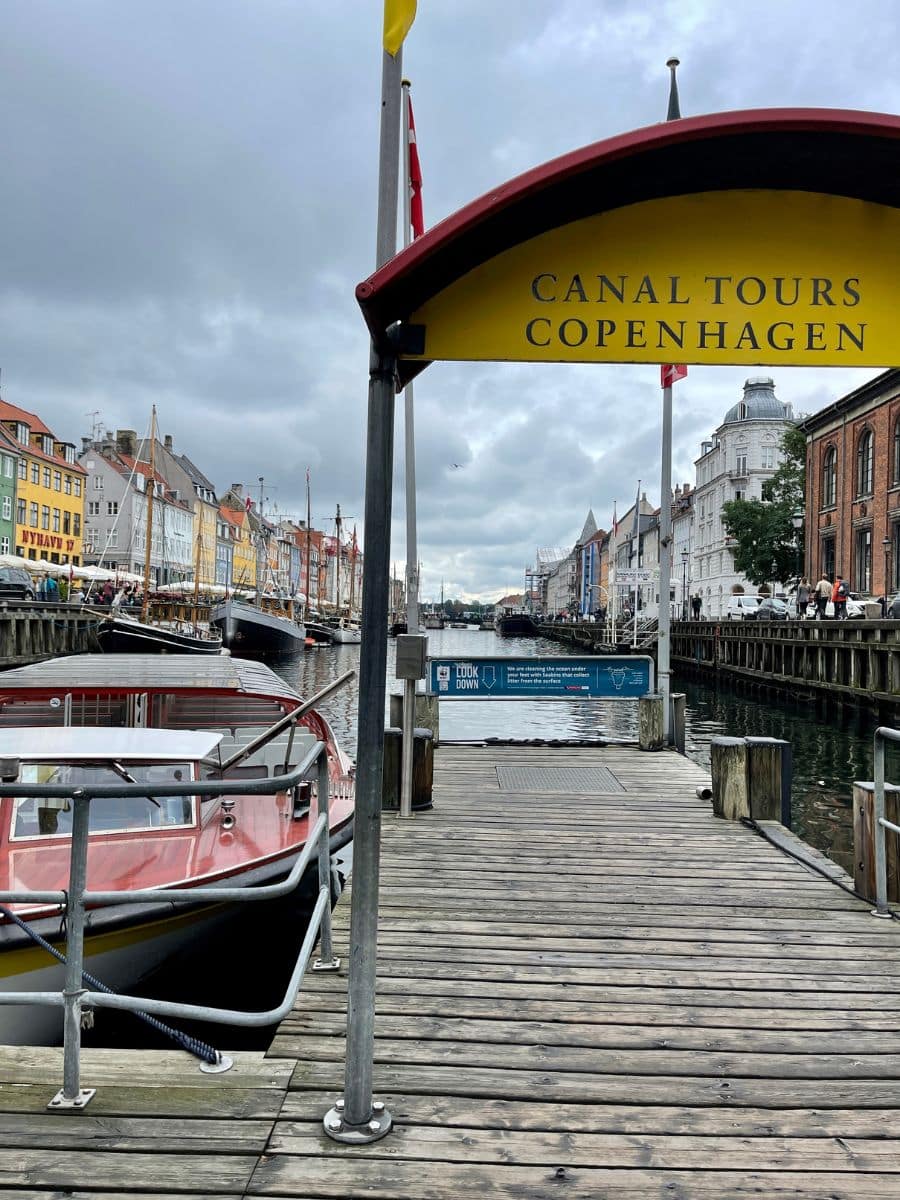 A wooden dock at Nyhavn, Copenhagen, with a yellow 'Canal Tours Copenhagen' sign arching overhead. A red and white canal tour boat is moored at the pier, ready to take passengers on a scenic waterway journey. The background showcases the iconic colorful buildings lining the harbor, with sailboats docked along the quay under an overcast sky.