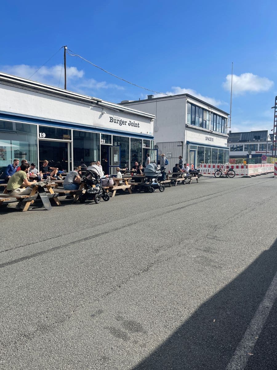 A casual outdoor dining scene at a burger joint in Copenhagen. People are sitting at wooden picnic tables, enjoying meals under a bright blue sky. The restaurant has a modern, minimalist white facade, and the surrounding area has an urban-industrial vibe with bicycles parked nearby and a construction barrier in the background.