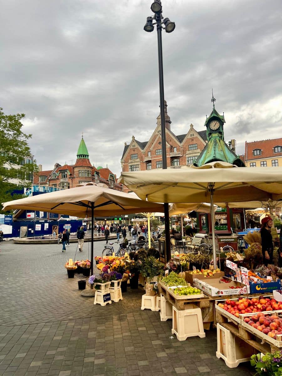 A vibrant outdoor market in Copenhagen with fresh fruits, vegetables, and flowers displayed under large beige umbrellas. The cobblestone plaza is surrounded by historic European-style buildings with green rooftops and intricate brickwork. A small kiosk with a green roof and clock is visible in the background, adding to the charm of the bustling city square. People are seen walking, shopping, and enjoying the lively atmosphere.