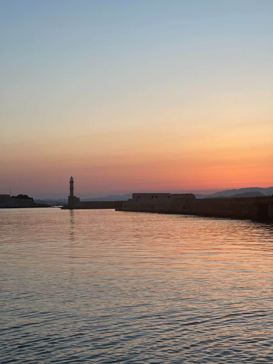 Sunset in Crete with a serene view of calm waters and a clear sky gradient from deep blue to orange. A lighthouse stands in the distance. This picturesque setting is a highlight for participants of an all-girls group tour exploring the beauty of Crete.