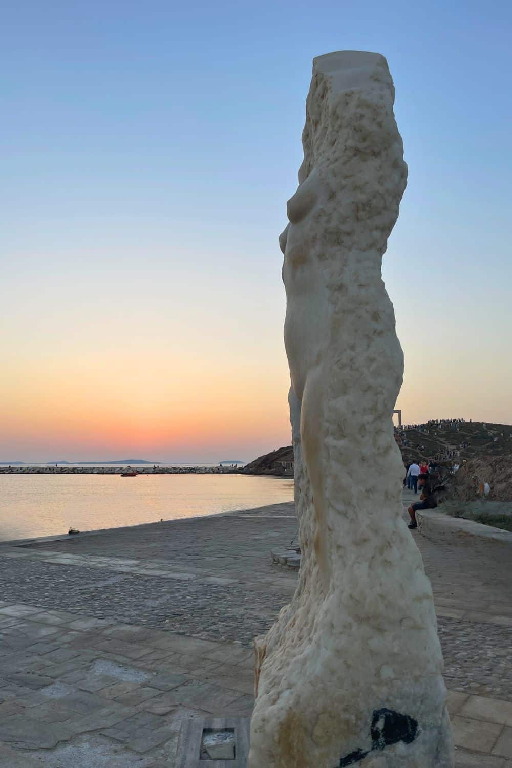 Silhouette of a modern, abstract sculpture resembling a standing figure against the backdrop of a soft sunset sky, with the calm sea stretching into the horizon and people milling about on the waterfront