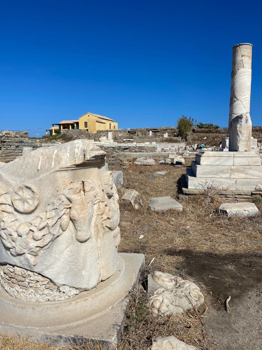 Close-up of ancient ruins on Delos, Greece, showing a carved marble fragment decorated with wreath designs in the foreground. A partially standing column rises on a pedestal nearby, with scattered stone remnants and a yellow building in the background under a clear blue sky.