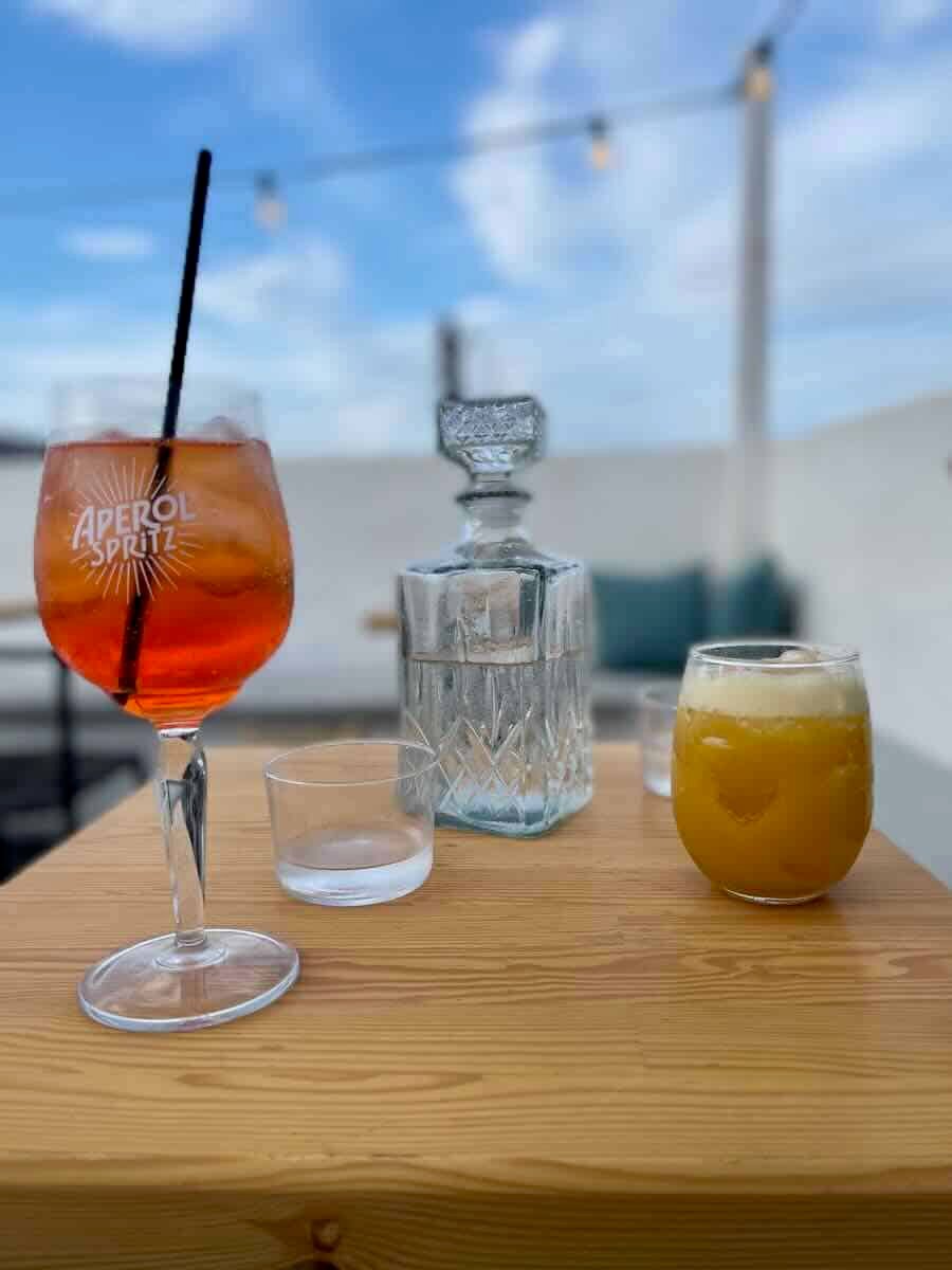The image shows an Aperol Spritz and a frothy orange drink on a wooden table, along with a crystal water decanter. The setting is outdoors with a clear blue sky and soft lighting, creating a relaxed vibe.