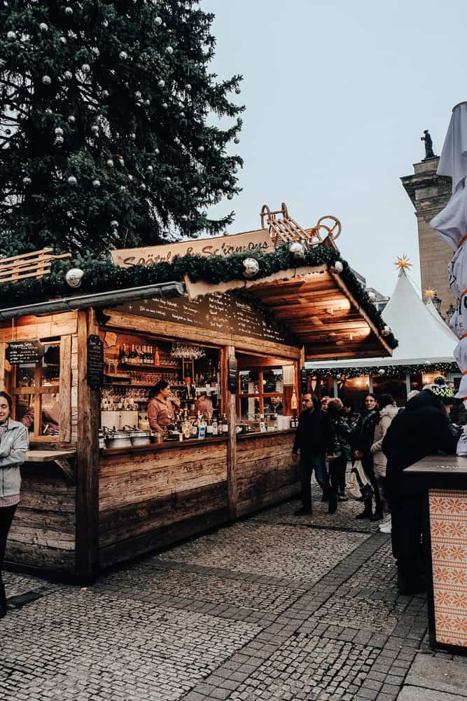 A bustling Christmas market scene with a wooden stall decorated with garlands and ornaments. People are gathered around, ordering food and drinks. A large, decorated Christmas tree and a historic building are visible in the background, adding to the festive atmosphere.
