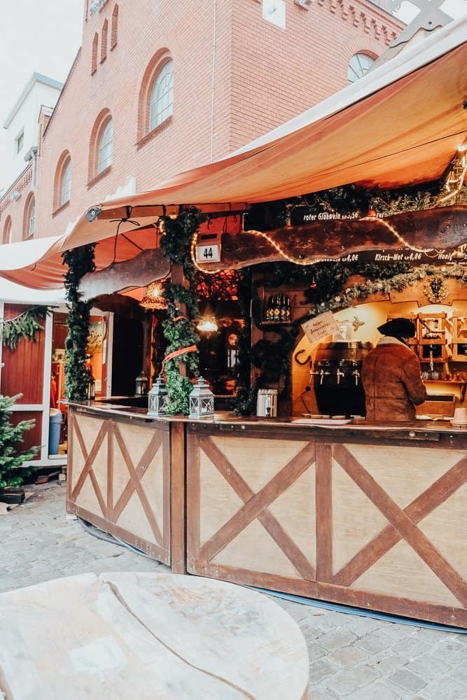 A quaint Christmas market stall in Berlin, adorned with festive greenery and lights, nestled against a historic building.