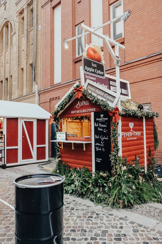 A Christmas market stall at Lucia Berlin Xmas Market, uniquely decorated with a large tomato and signs for hot drinks, nestled between traditional buildings.
