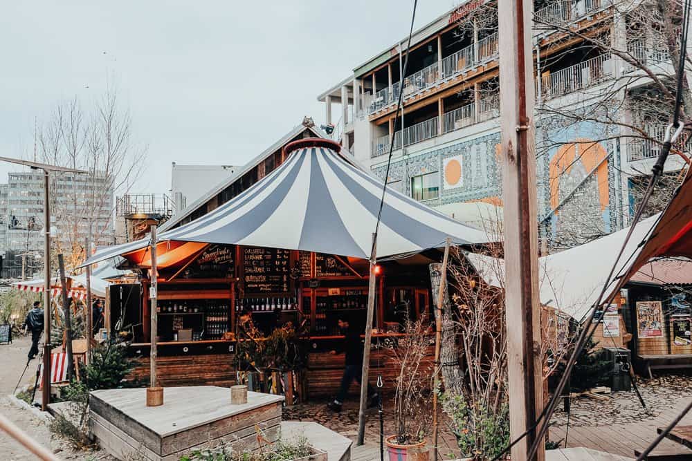 A warm and inviting wooden stall at a Berlin Christmas market, sheltered under a large canopy, surrounded by urban art and architectural elements.