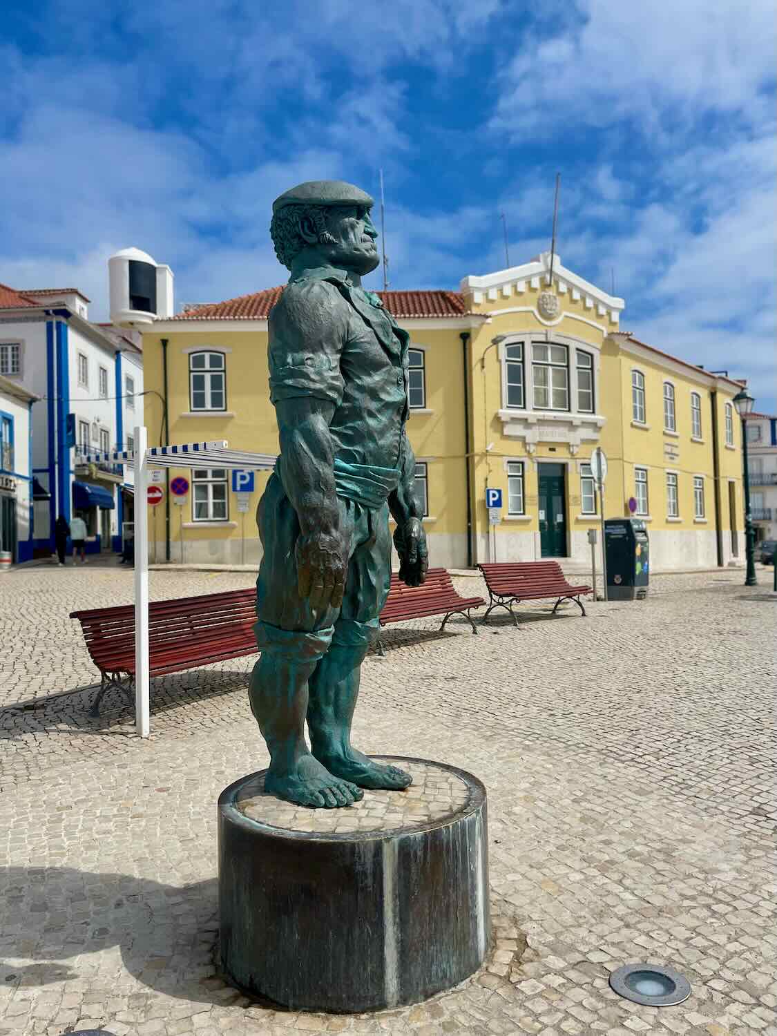 A bronze statue of a fisherman stands proudly in a square in Ericeira, Portugal. The figure, dressed in traditional attire, captures the town’s deep connection to the sea and its fishing heritage. Behind the statue, colorful benches and a charming yellow building with red roof tiles add to the picturesque and coastal charm of Ericeira, with a bright sky dotted with clouds overhead.