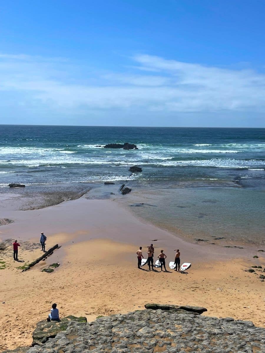 A scenic beach in Ericeira, Portugal, with golden sand, rocky formations, and rolling ocean waves. A group of young surfers in wetsuits stands near the shoreline with their surfboards, preparing to enter the water. Other beachgoers are scattered along the shore, some walking, others sitting on the rocks, enjoying the view of the deep blue Atlantic Ocean under a bright sky with soft clouds."