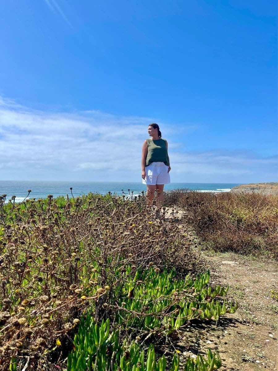 Melissa stands on a coastal cliff in Ericeira, Portugal, gazing into the distance. She wears a green sleeveless top and white shorts, with her hair pulled back. The foreground features dry and green coastal vegetation, while the ocean stretches out behind her. The sky is bright blue with wispy clouds, creating a serene and picturesque scene.