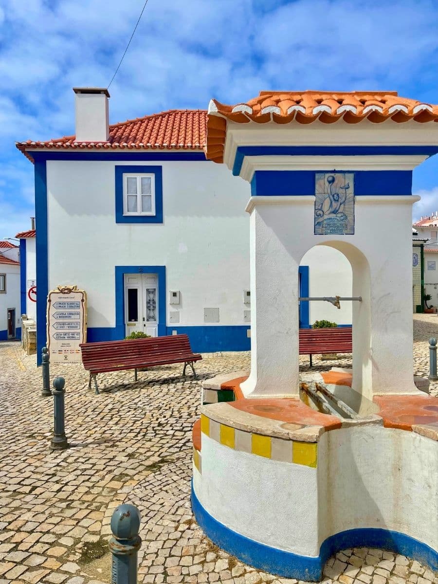 A bright, sunlit village square with cobblestone paving, a traditional whitewashed fountain topped with terracotta tiles and accented in blue and yellow. In the background, a charming white house with a red tiled roof and blue trim around the windows and doors. Two red benches sit along the path, inviting rest under a clear blue sky.