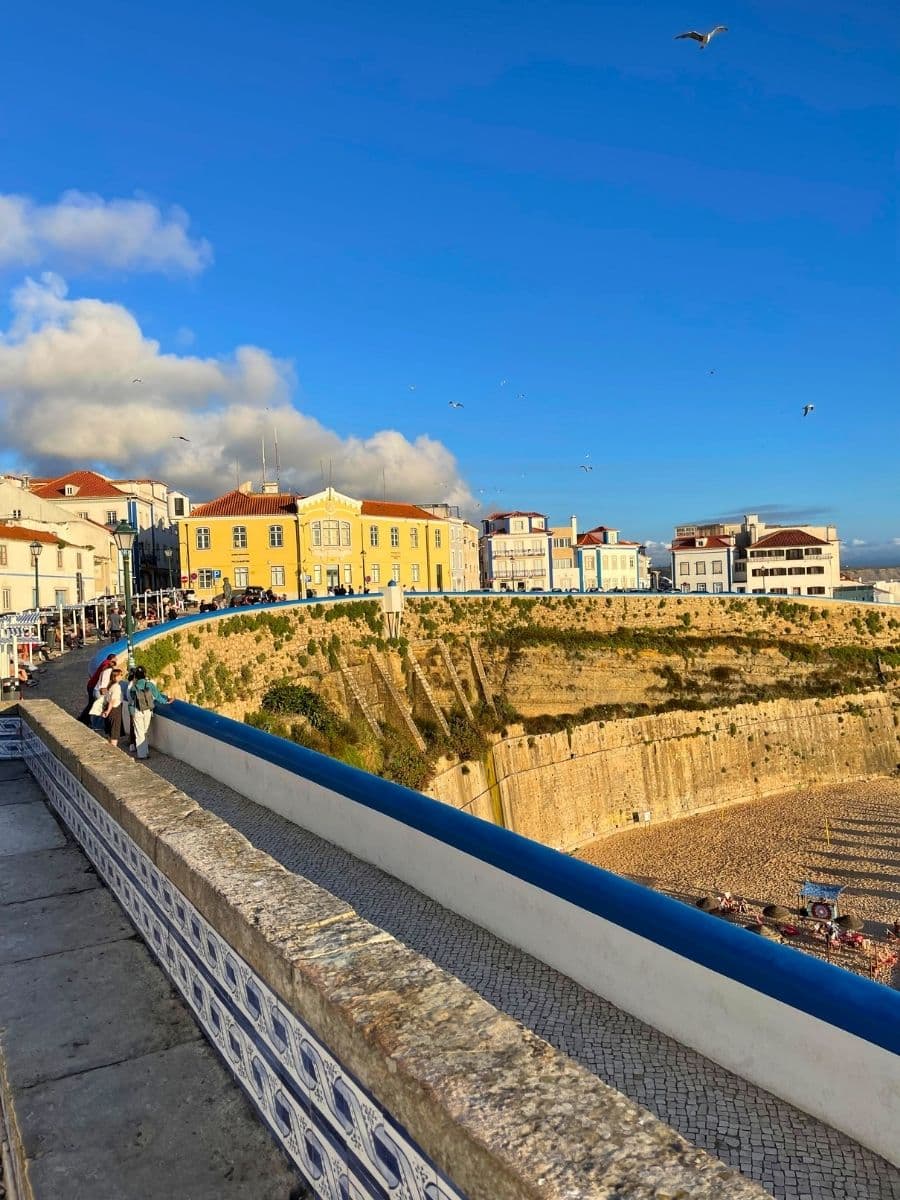 A picturesque view of a coastal promenade in Ericeira, Portugal, overlooking a sandy beach below. The stone walkway is lined with traditional Portuguese tiles, and a blue and white railing separates pedestrians from the steep cliffside. People are seen strolling along the path, enjoying the sunny day. In the background, colorful buildings with red-tiled roofs contrast against the deep blue sky, while seagulls soar above, adding to the charm of this seaside town.