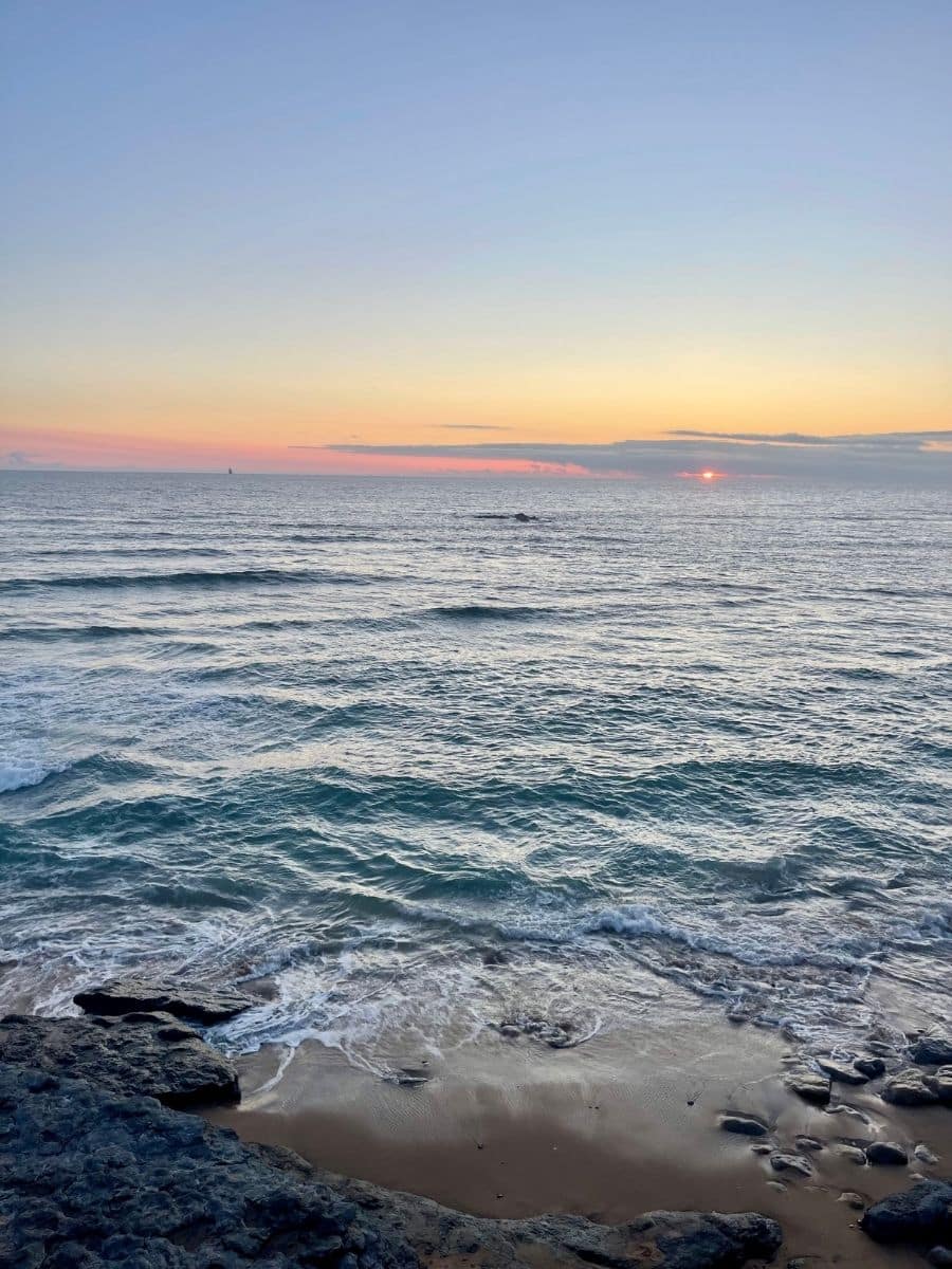 A serene coastal scene in Ericeira, Portugal, captures the sun setting over the Atlantic Ocean. Gentle waves roll onto a sandy beach, their motion creating delicate white froth as they meet scattered dark rocks. The sky displays a gradient of soft pastels, with warm oranges and pinks blending into the cool blues of the evening. The horizon stretches far into the distance, where a lone sailboat is faintly visible. The peaceful atmosphere, combined with the rhythmic sounds of the waves, evokes a sense of calm and reflection.