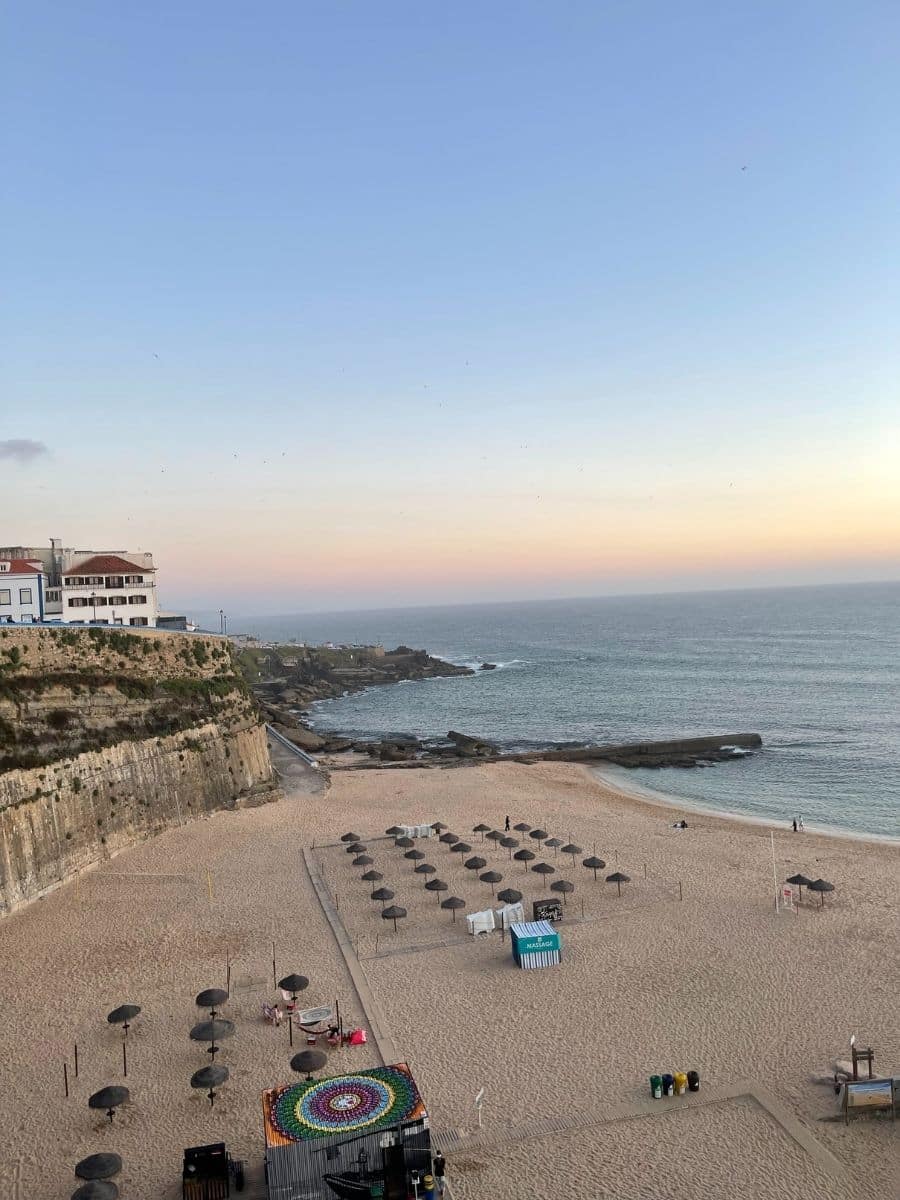 A serene coastal view at sunset featuring a sandy beach lined with neatly arranged straw umbrellas. A colorful, circular mosaic decorates a structure near the foreground. The beach extends towards rocky cliffs on the left, with a cluster of white buildings perched above. The ocean gently meets the shore, and a few people can be seen strolling along the water's edge. The sky transitions from a soft blue to warm pastel hues as the sun sets, casting a peaceful glow over the landscape.