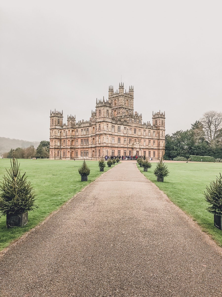 The image shows Highclere Castle at the end of a long pathway, surrounded by neatly trimmed lawns and potted shrubs. The overcast sky adds to the grand, serene atmosphere of the historic building.






