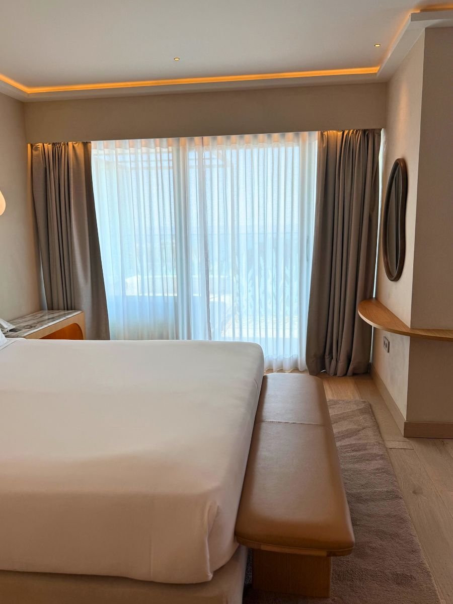 Bed facing full-length curtains with soft daylight streaming in, a leather bench at the foot of the bed, and a round mirror on the side wall.