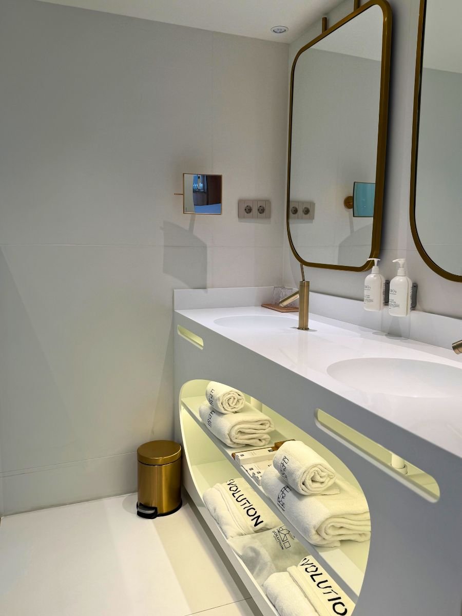 Modern double sink vanity with backlit shelving displaying neatly folded towels, gold fixtures, and a rectangular mirror.