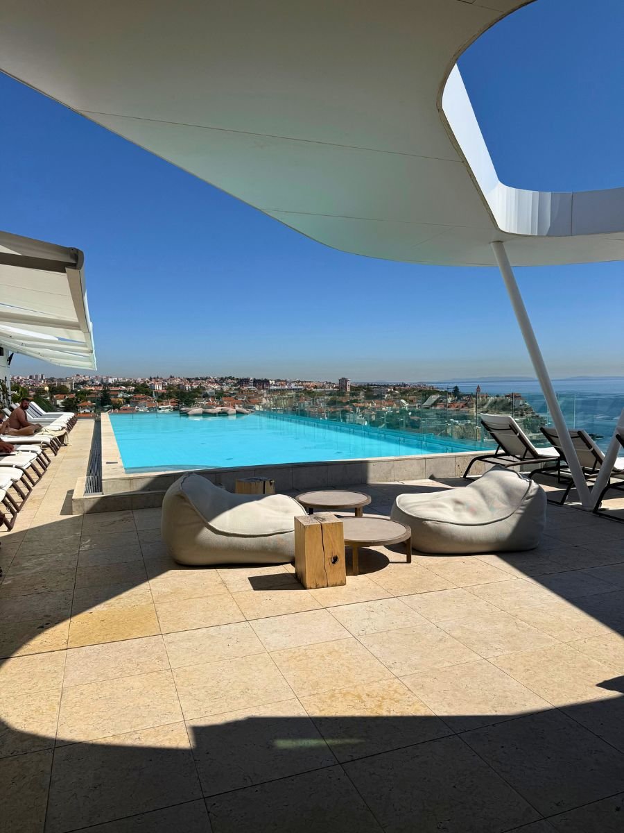 Rooftop infinity pool overlooking the city and coastline, with lounge chairs and beanbag seating under a canopy.