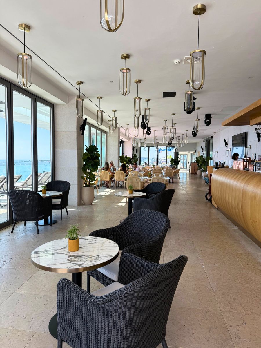 Bright dining space with marble tables, wicker chairs, and large windows overlooking the ocean.