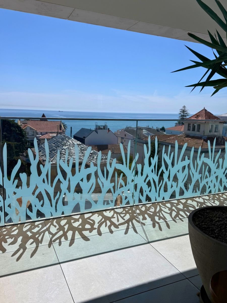 Glass balcony railing decorated with a coral-like design, overlooking rooftops and the ocean under a clear sky.