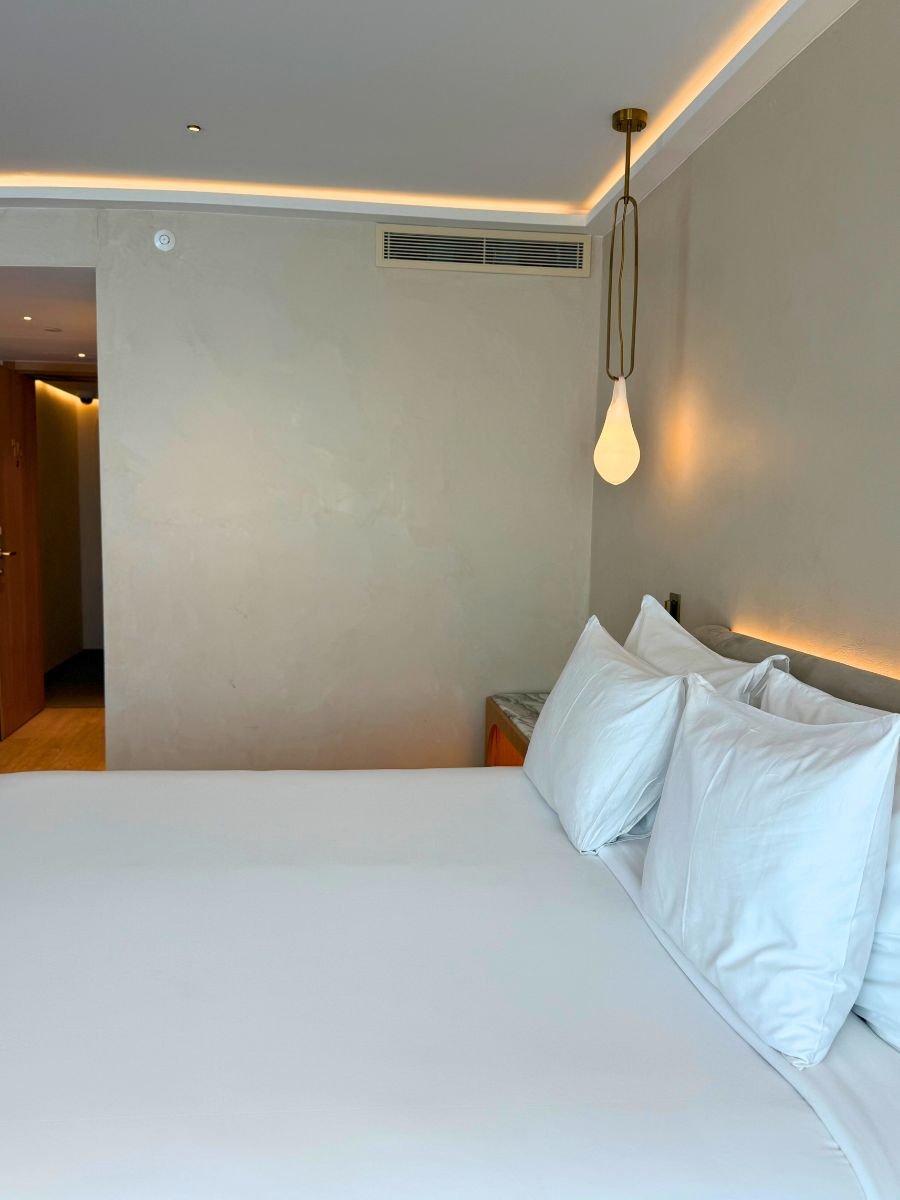 Crisp white bed with three pillows, marble nightstand, pendant light, and subtle ceiling lighting in a minimalist hotel room.