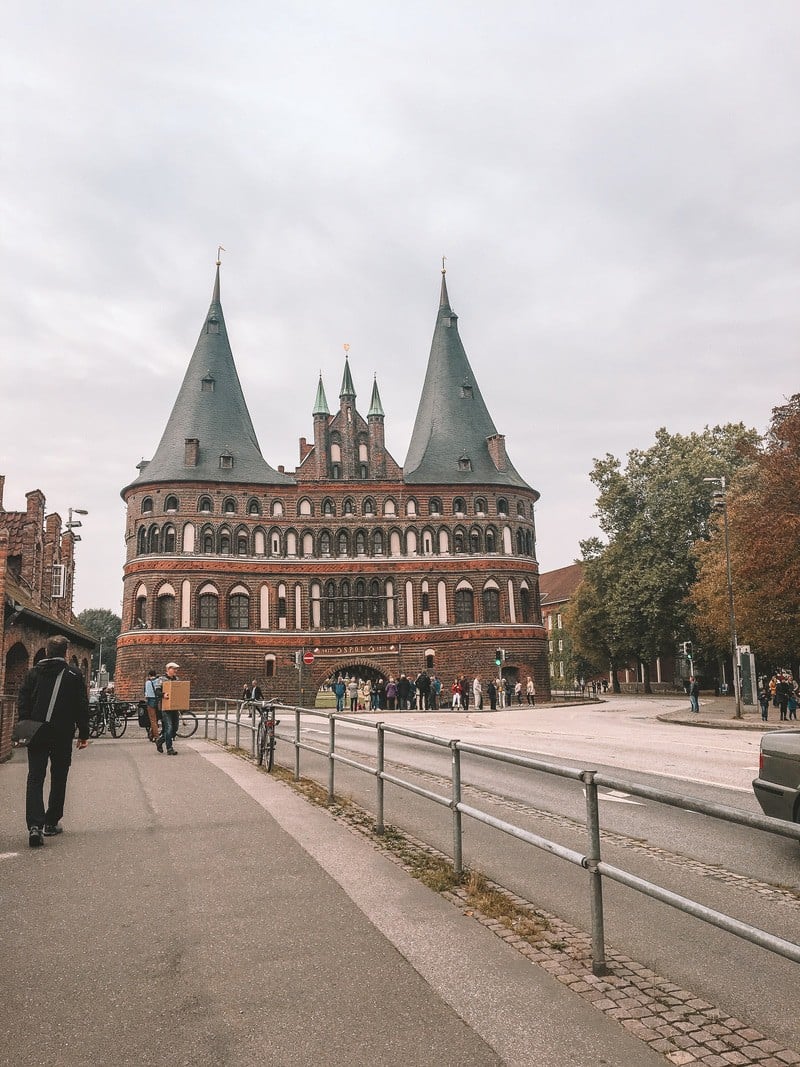 A historic, medieval gate with two large, conical towers stands prominently in the center of the image. The structure, made of dark brick with lighter accents, features multiple arched windows and a small, central archway leading through it. The scene is set on a paved street with a few pedestrians and cyclists, with trees and other buildings visible in the background under an overcast sky. The architecture exudes a sense of history and old-world charm.