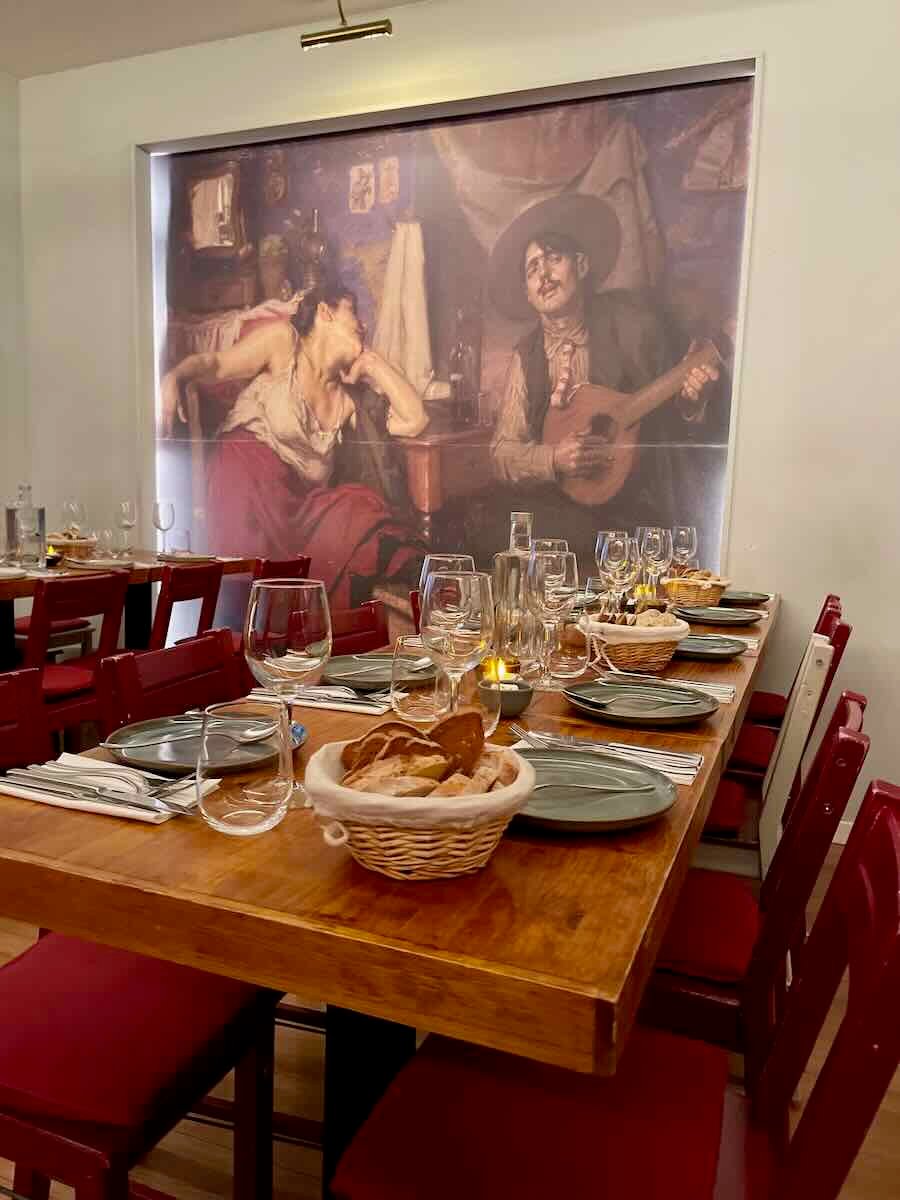 A long wooden dining table is elegantly set with wine glasses, plates, and bread baskets. Behind the table, a large mural of a man playing a guitar and a woman leaning on her arm creates an intimate, artistic atmosphere. The room features red chairs and a warm, welcoming vibe.