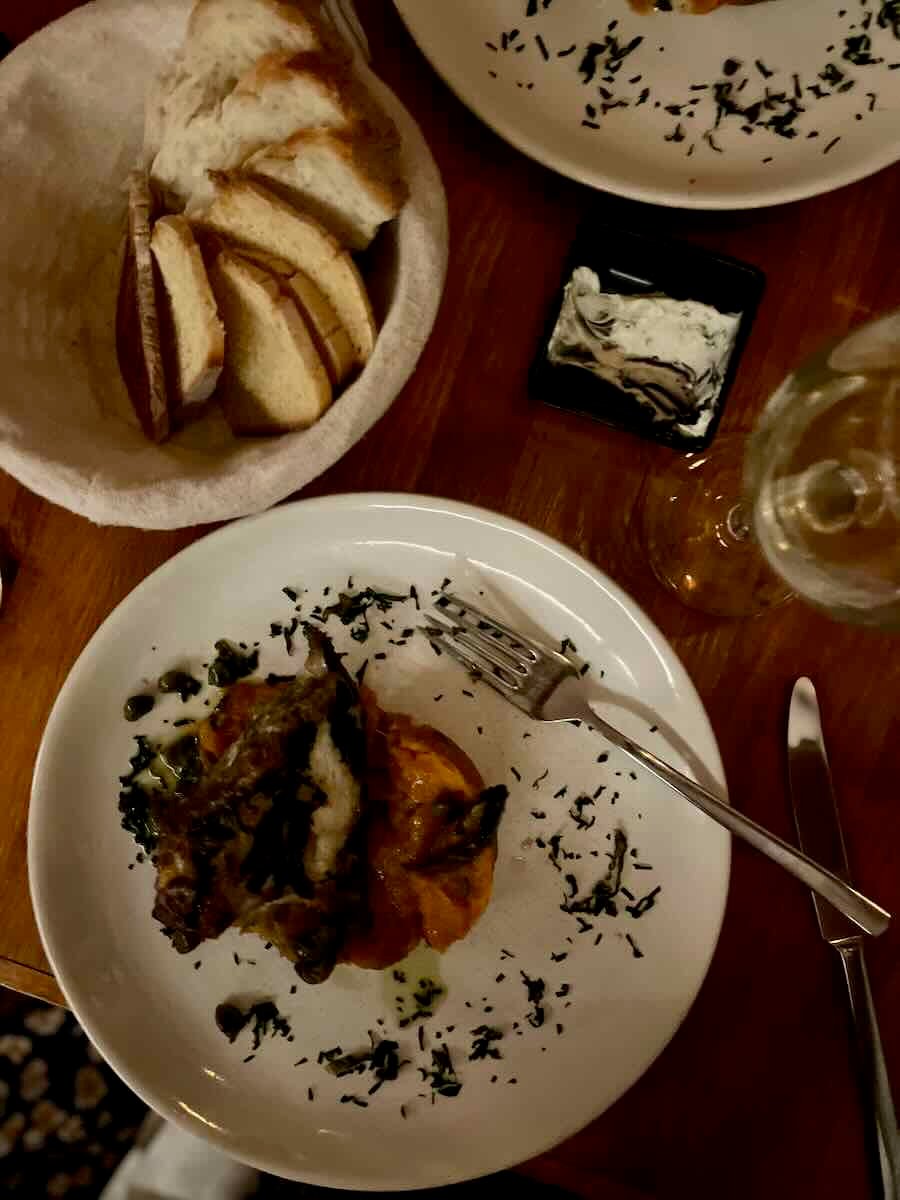 A dinner plate with a carefully plated dish of meat topped with vegetables, surrounded by a light drizzle of herbs. Next to the plate is a basket of sliced bread, a small dish of spreadable butter, and a glass of wine on a wooden table, set for a cozy meal.